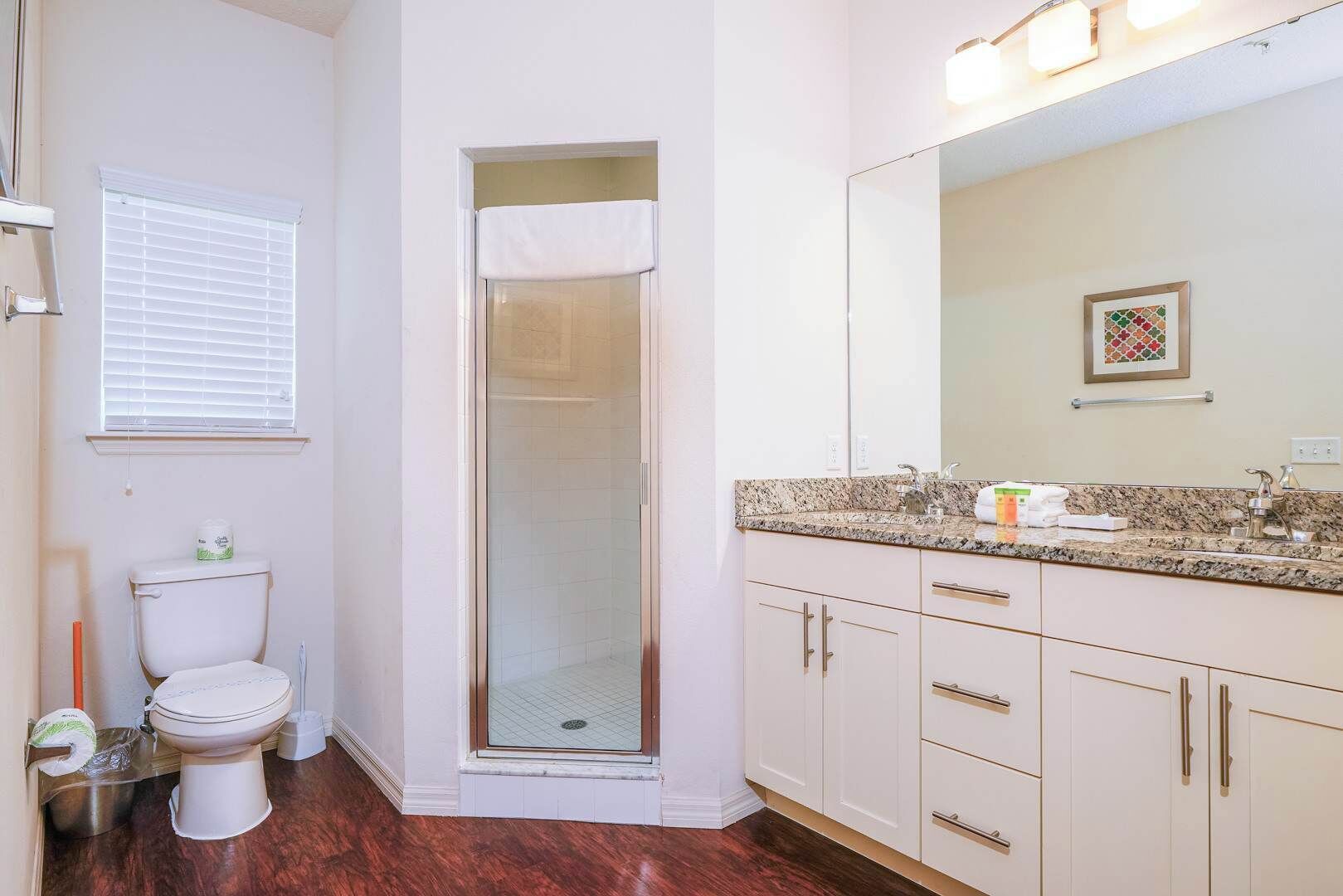 A bathroom with a toilet, shower, vanity with a mirror, and a window with blinds.