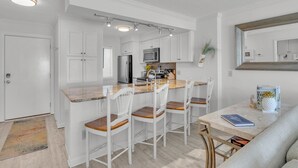 A modern kitchen with white cabinetry, a central island with seating, and a dining area with a table and chairs. - Seaside Vista, Luxury Oceanfront, Top "2nd" Floor, Amazing Views!! (Kure Beach)
