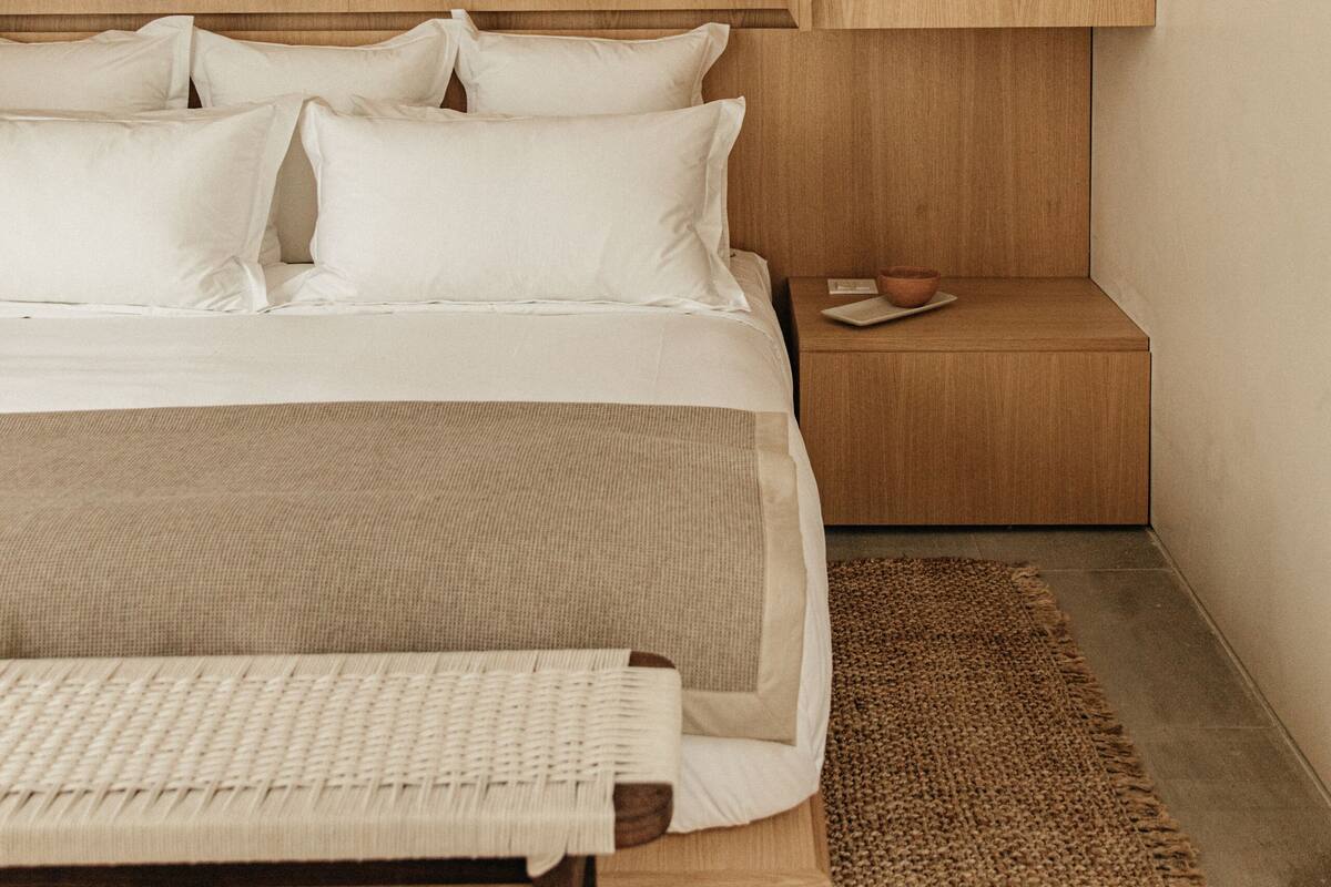 A modern bedroom with a wooden headboard, a bed with white linens, a bedside table with a bowl, and a woven rug.
