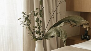 Une chambre à coucher avec un lit, une table de chevet ornée d’un vase blanc et d’une plante, un banc en osier et une tête de lit en bois.