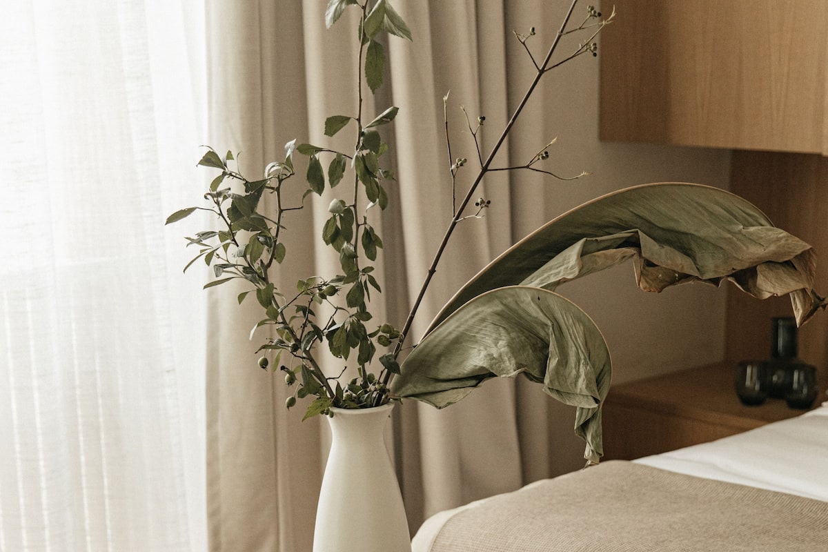 A bedroom with a bed, a bedside table with a white vase and a plant, a woven bench, and a wooden headboard.