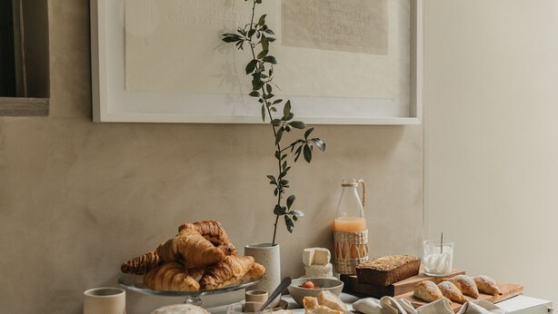 Une table de petit-déjeuner avec des viennoiseries, du pain, du fromage et une bouteille de jus d’orange.