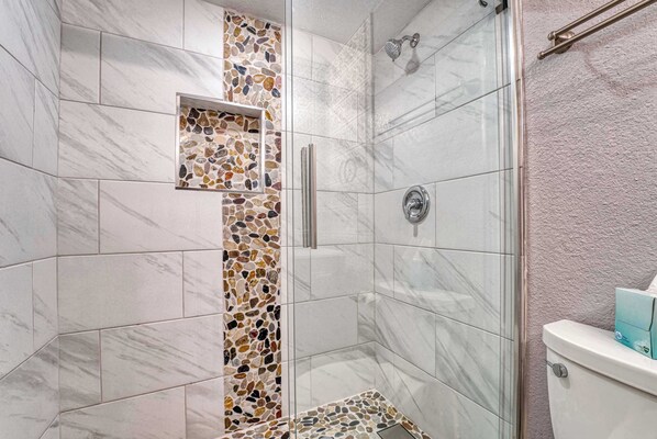A shower with a glass door, a tiled wall with a mosaic pattern, and a toilet with a blue tissue box on it.
