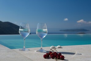 Dos vasos de agua y un racimo de cerezas sobre una mesa junto a una piscina con vistas al océano y las montañas.