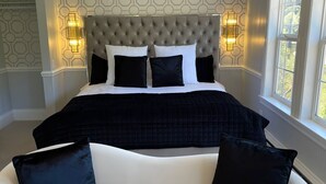 Un dormitorio con cabecera capitonada, una cama grande con ropa de cama blanco y negro, dos lámparas fijadas a la pared y un sofá blanco con cojines negros.