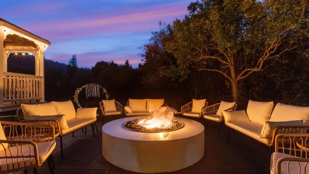 Un espacio de estar al aire libre con forma circular, que incluye una chimenea, sillas de mimbre y una estructura blanca al fondo.