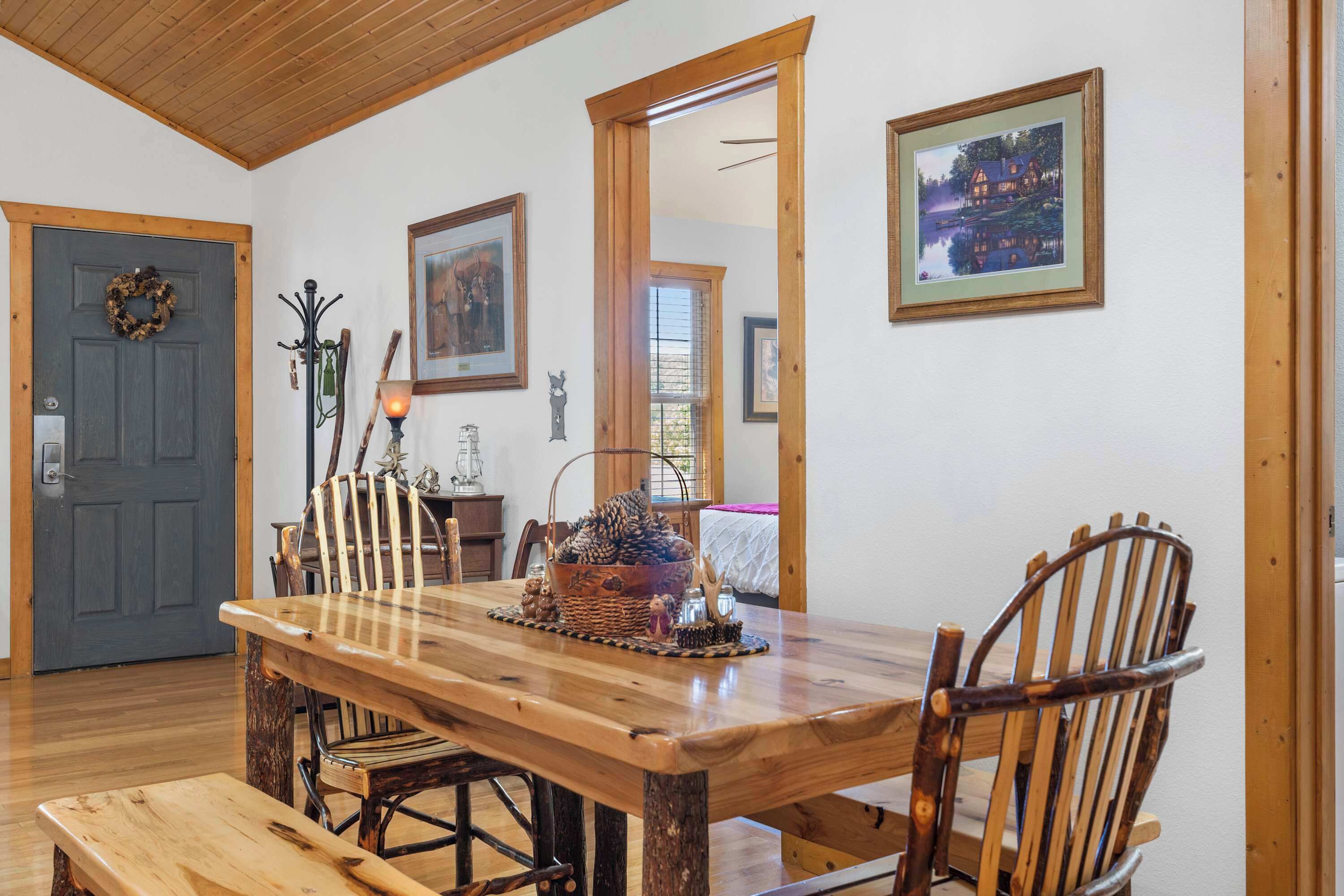 Un área de comedor con una mesa y sillas de madera, un banco, una cesta con piñas y un cuadro en la pared.