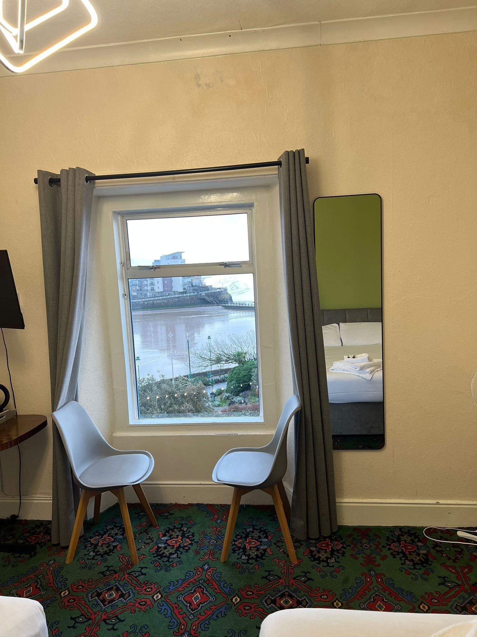 A hotel room with a large window, two chairs, a bed, a mirror, and a patterned carpet.