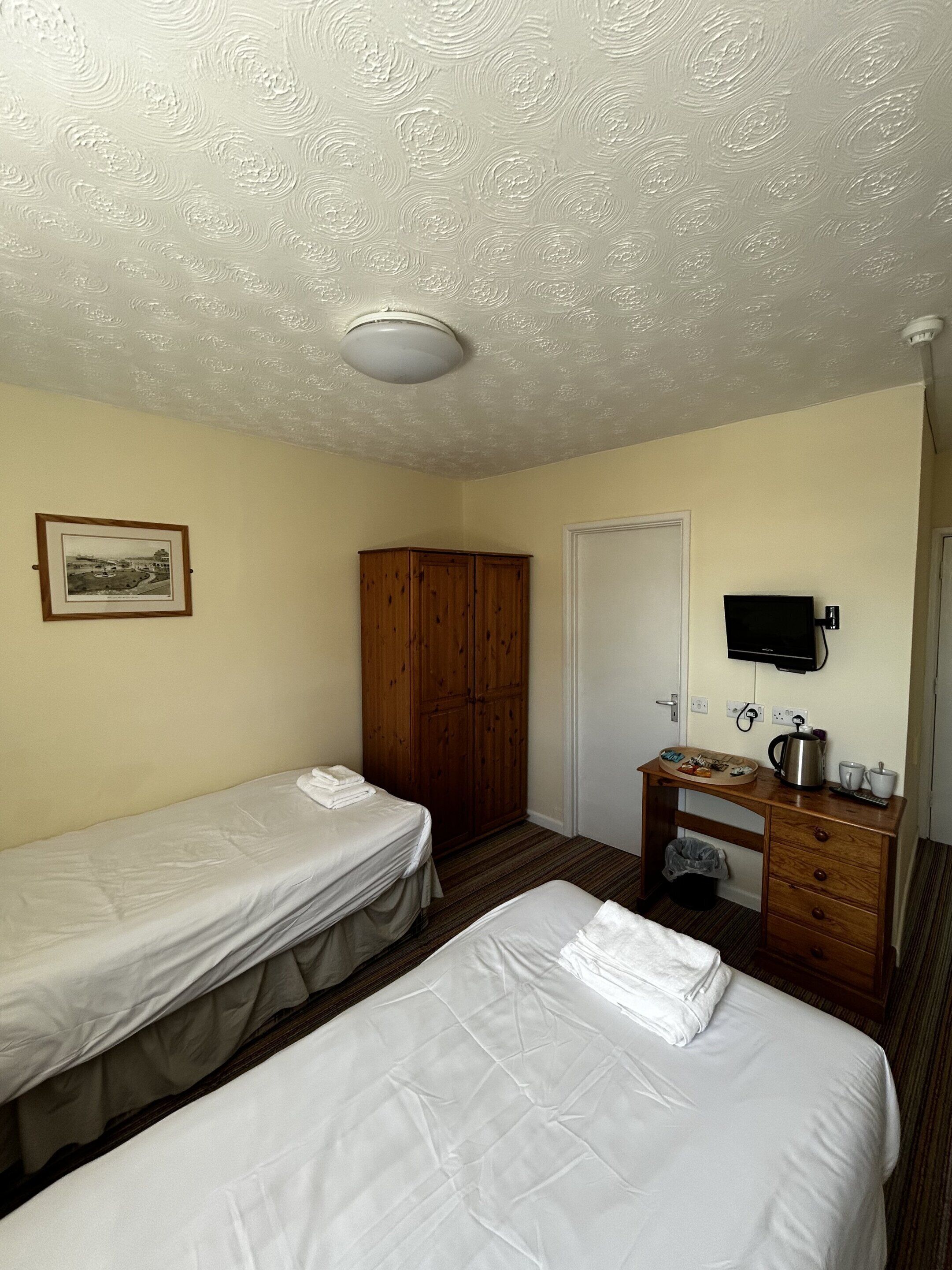 A hotel room with two single beds, a wooden wardrobe, a desk with a kettle, cups, and a remote control.