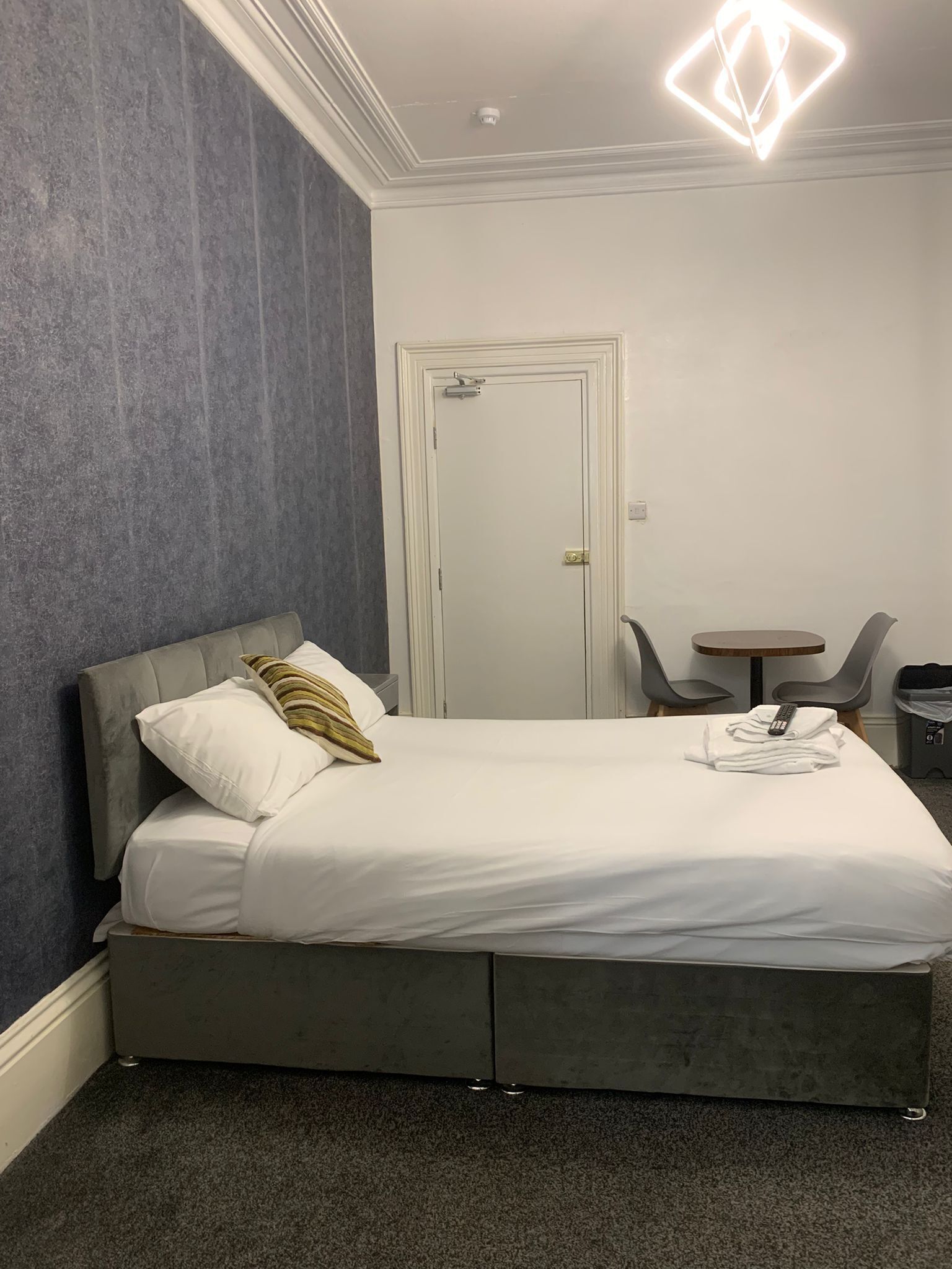 A modern hotel room with a grey bed, white bedding, a grey headboard, a small round table with two chairs, and a door with a handle.
