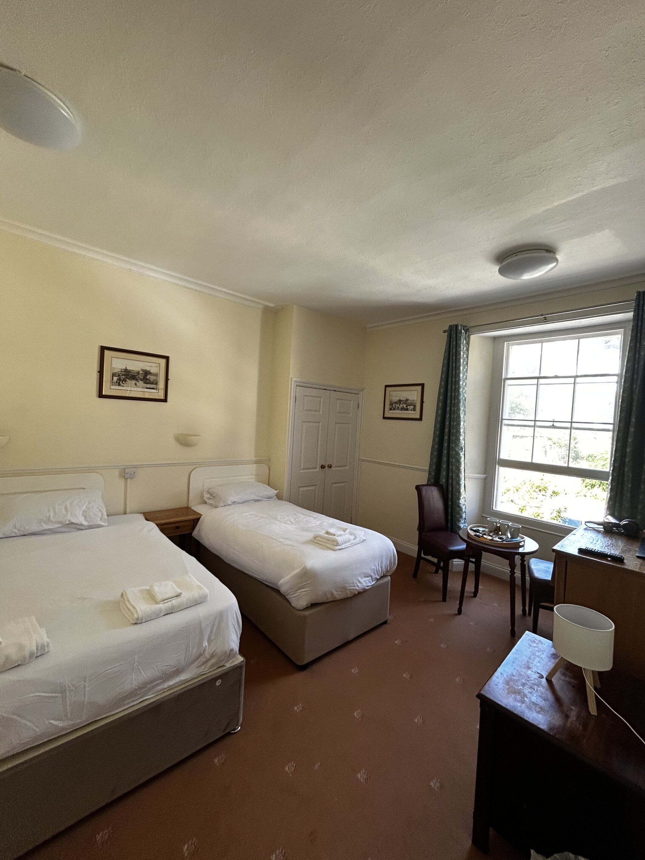 A hotel room with two beds, a desk with a chair, a window with curtains, and a door.