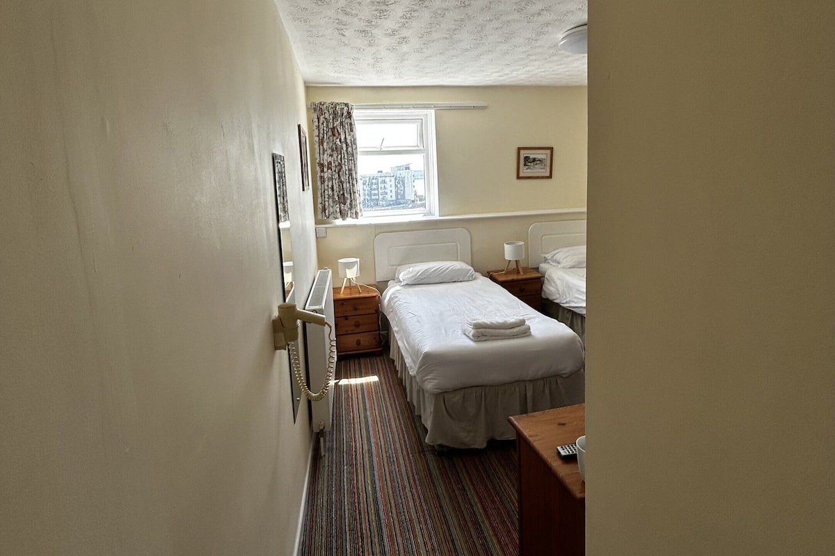 Une chambre d’hôtel avec un lit simple, une table de chevet, une commode, une fenêtre avec des rideaux et un radiateur.