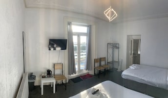 Habitación de hotel con cama, televisor montado en la pared, ventana con cortinas, radiador, una mesita con hervidor y tazas, y un perchero.