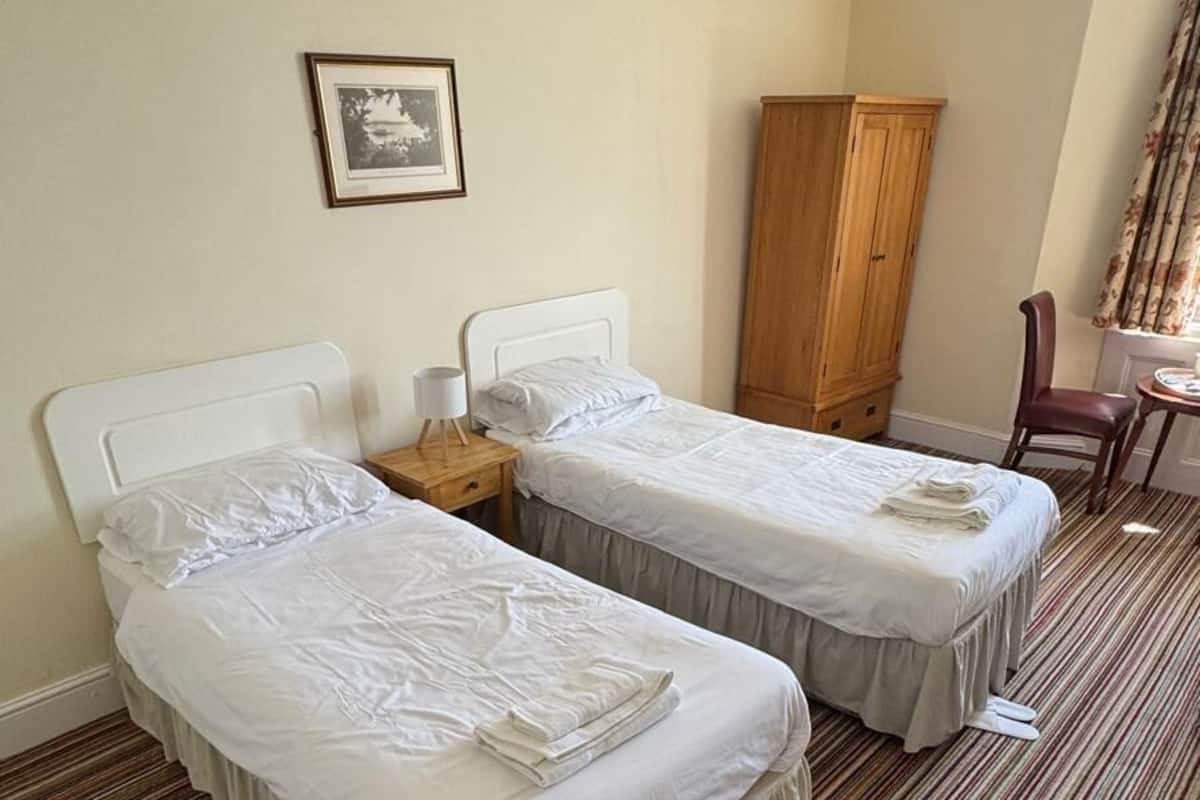 Une chambre d’hôtel avec deux lits simples, une armoire en bois, une chaise et un tableau encadré au mur.