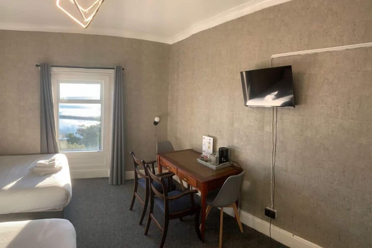 Une chambre d’hôtel comprenant un lit, un bureau avec des chaises, un téléviseur fixé au mur et une fenêtre avec des rideaux.