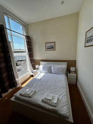 A hotel room with a bed, two bedside tables, a window with curtains, a framed picture on the wall, and a view of the outside. - Savoy Hotel (Weston-super-Mare)