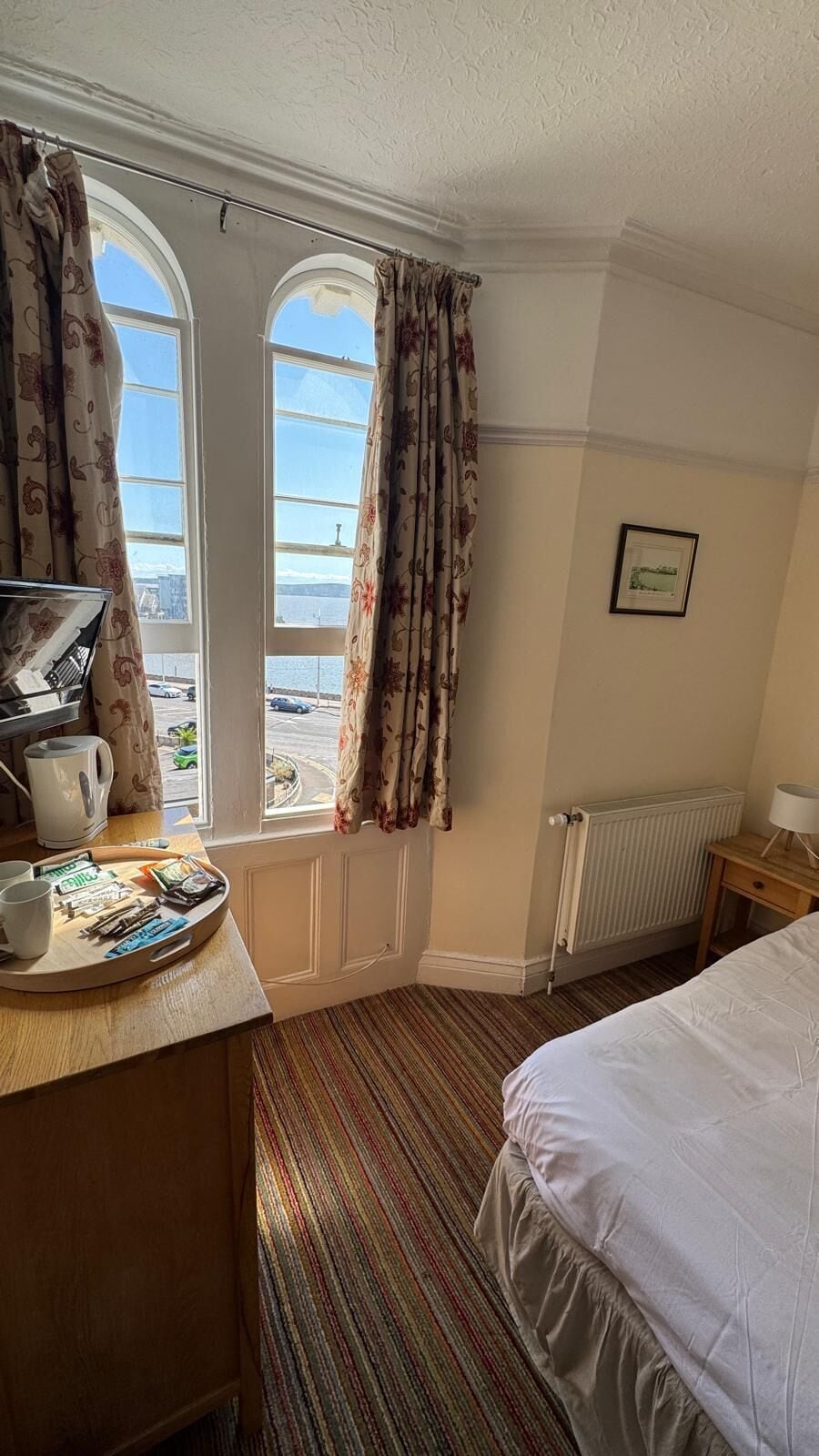 A hotel room with a bed, a desk with a tray of breakfast items, a radiator, and a window with curtains.