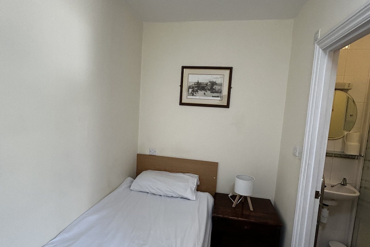 Une chambre d’hôtel avec un lit simple, une table de chevet, un tableau encadré au mur et une salle de bain visible à travers une porte ouverte.