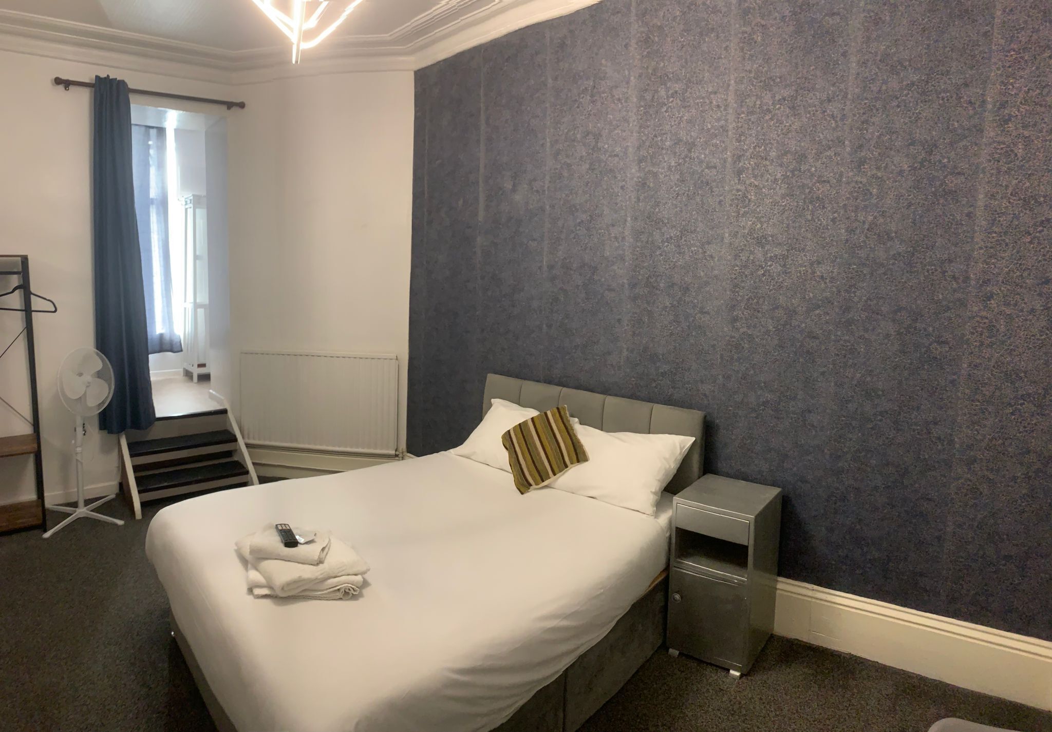 Habitación de hotel con cama, mesita de noche, ventilador y vista al exterior.
