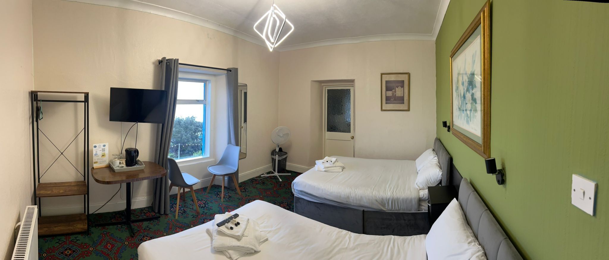 A hotel room with two beds, a desk with a flat-screen TV, a chair, a standing fan, and a window with curtains.