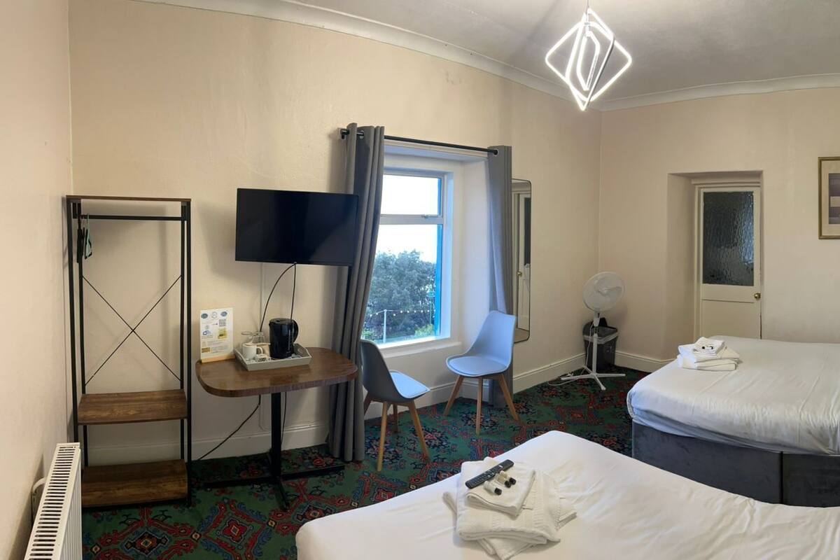 Quarto de hotel com duas camas, uma mesa de trabalho, televisão de tela plana, uma cadeira, ventoinha e janela com cortinas.