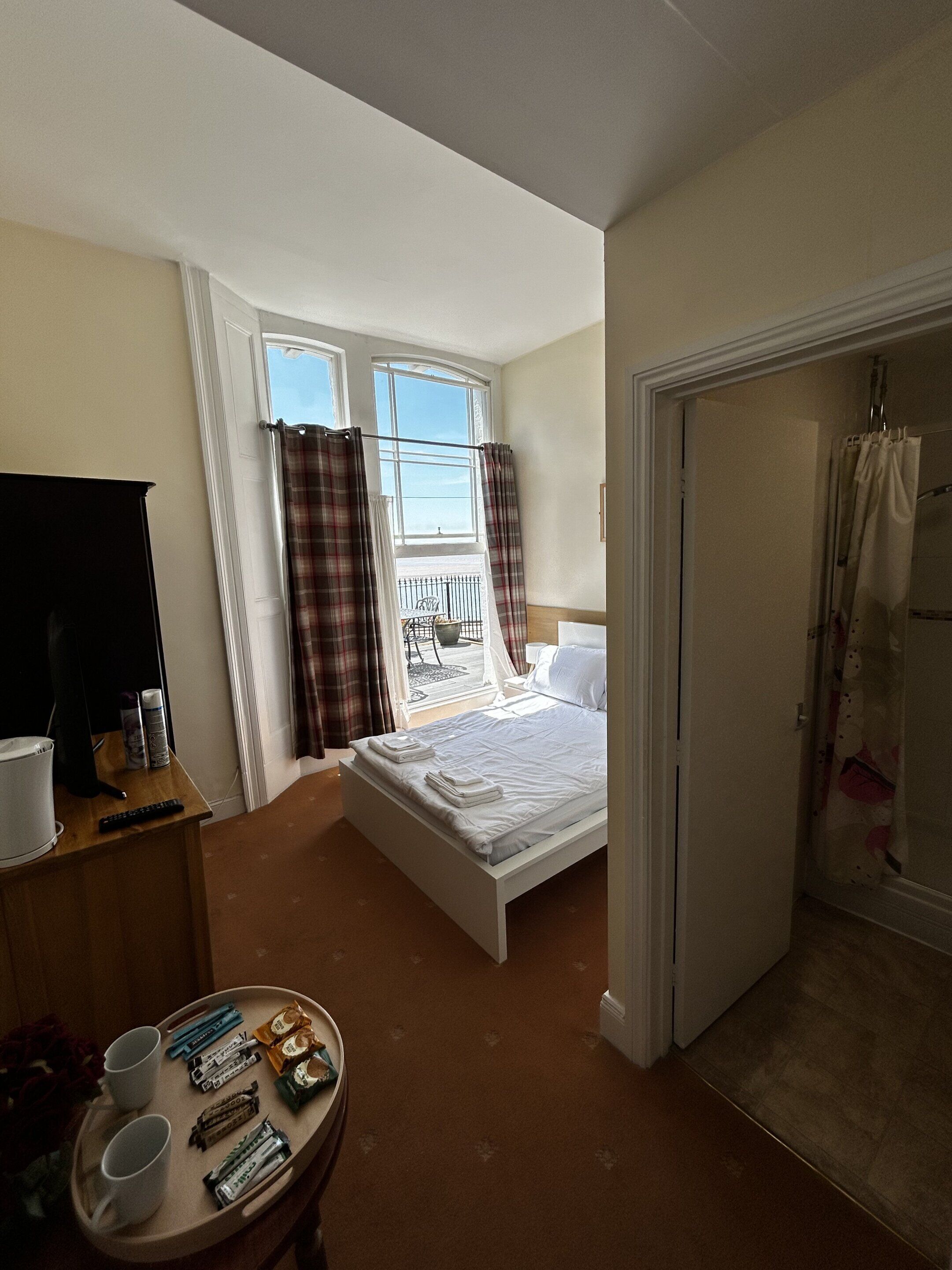Habitación de hotel con cama, escritorio con computadora y silla, una mesita con objetos, y ventana con cortinas.