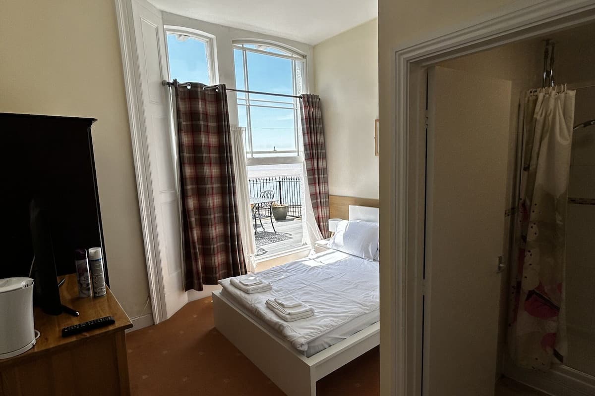 Une chambre d’hôtel comprenant un lit, un bureau avec un ordinateur et une chaise, une petite table avec quelques objets, et une fenêtre avec des rideaux.