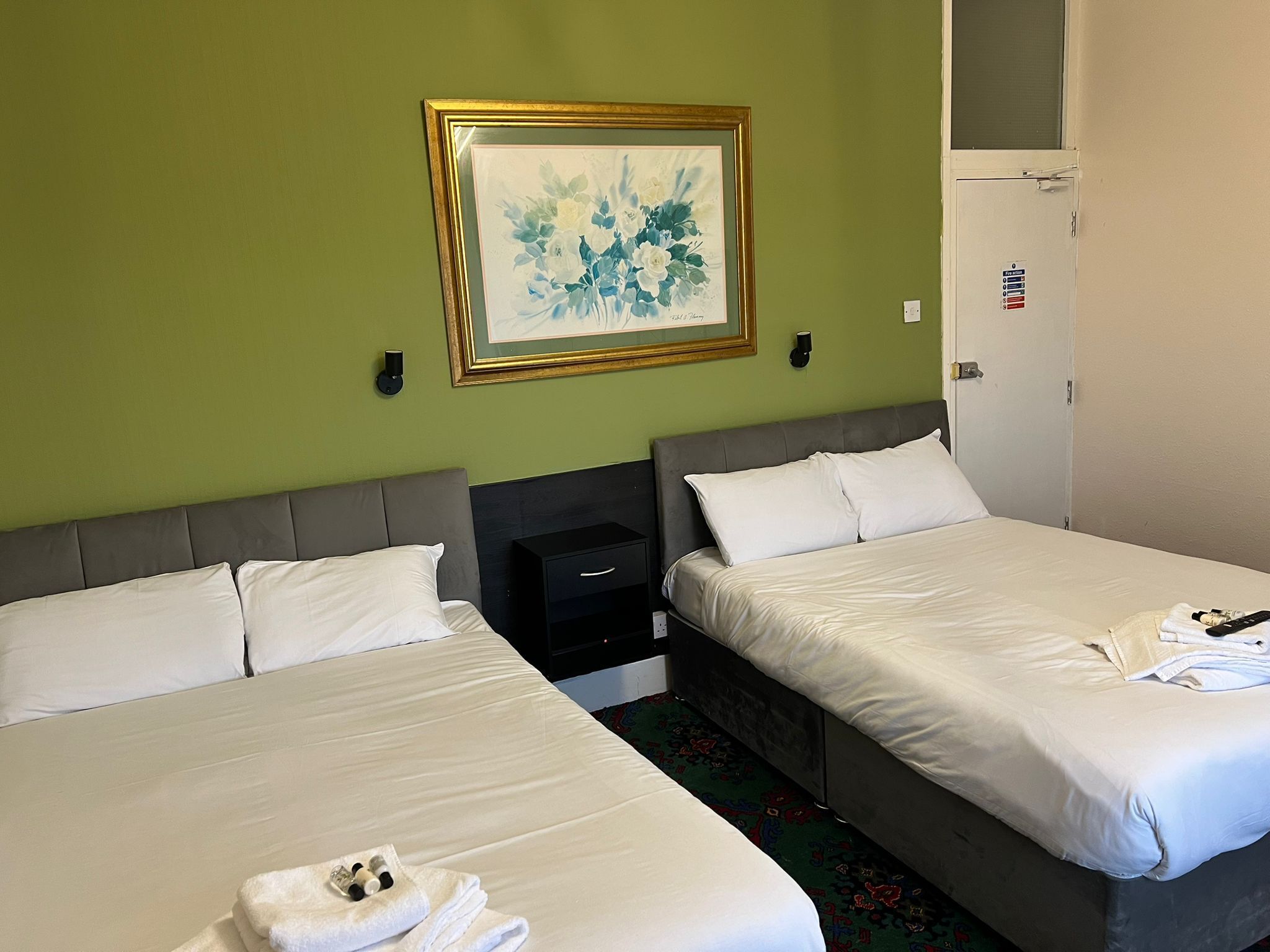 Una habitación de hotel con dos camas, un cuadro floral enmarcado en la pared, una mesita de noche negra pequeña y una puerta con un letrero de seguridad.