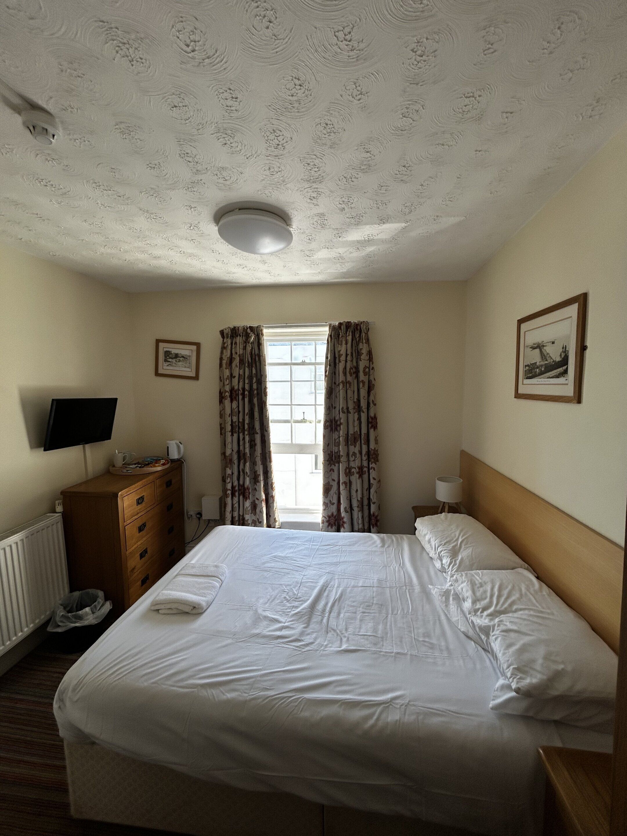 Habitación de hotel con cama, cómoda, televisión, ventana con cortinas e imagen en el marco en la pared.
