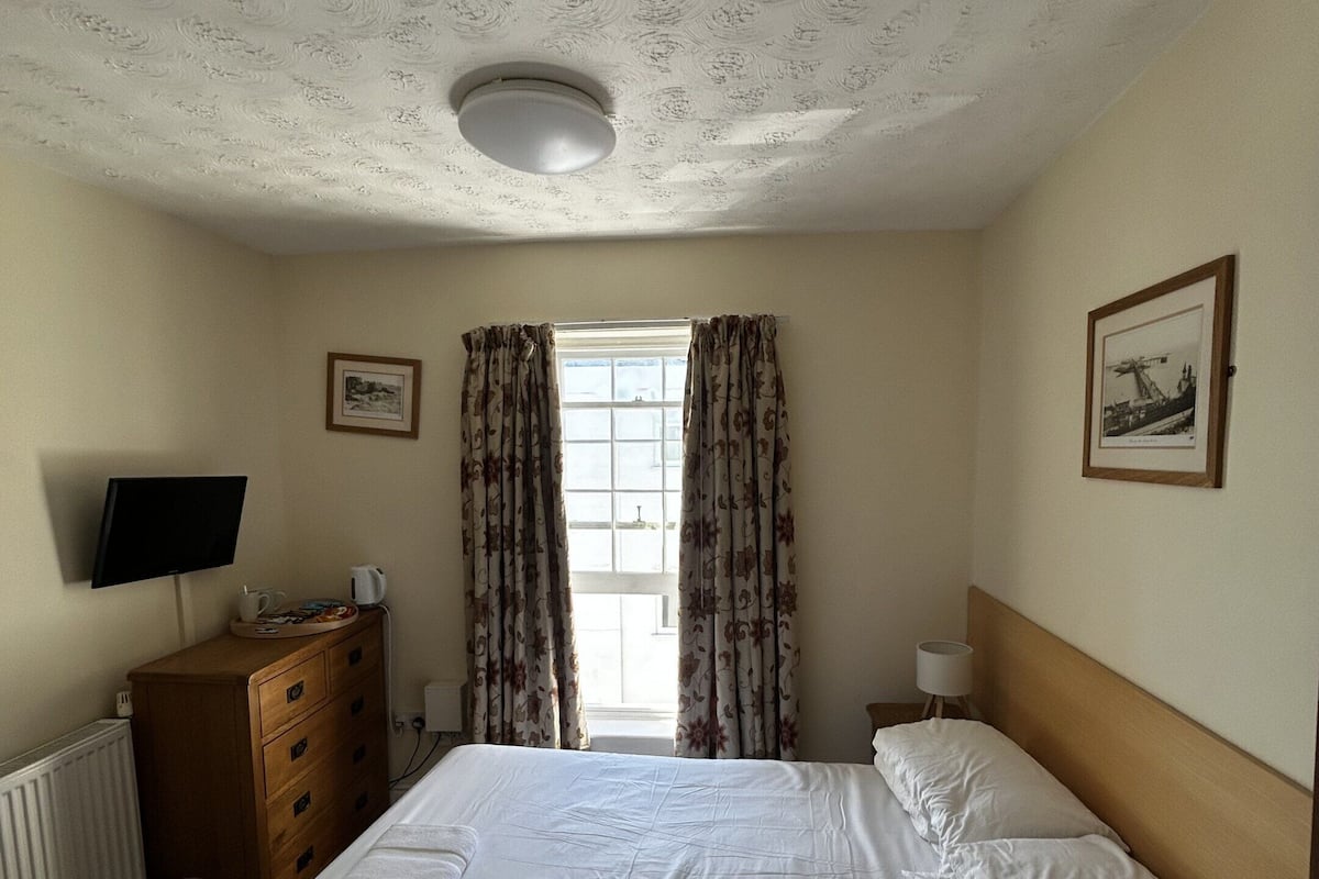Une chambre d’hôtel avec un lit, une commode, une télévision, une fenêtre avec des rideaux et un tableau encadré au mur.