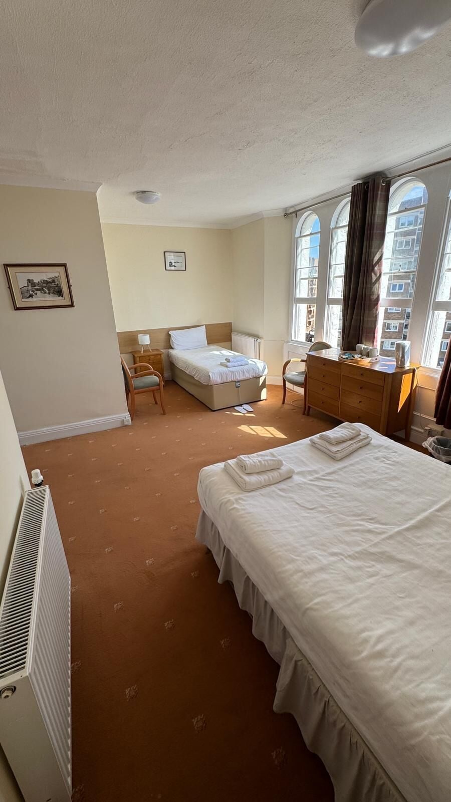 A hotel room with a bed, a desk, a chair, a radiator, and large windows.