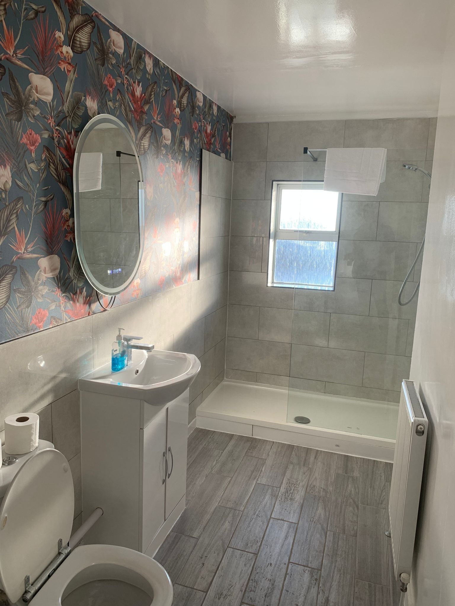A bathroom with a toilet, sink, mirror, shower, and a window.