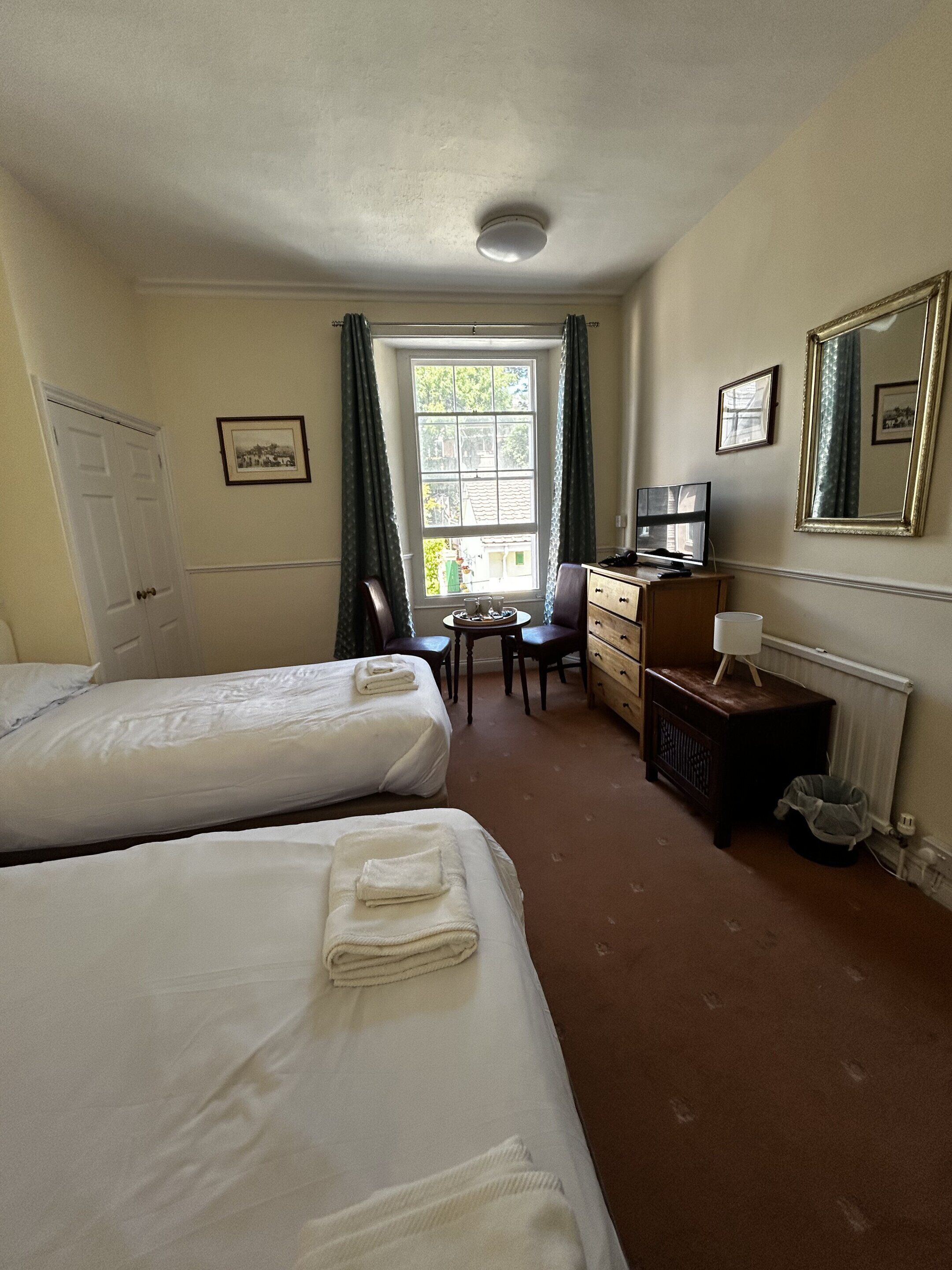Habitación de hotel con dos camas, un escritorio con silla, cómoda, radiador y ventana con cortinas.