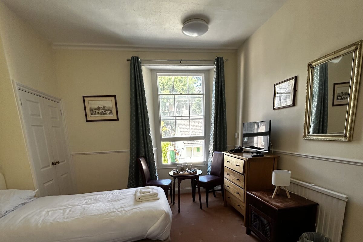 Une chambre d’hôtel avec deux lits, un bureau avec une chaise, une commode, un radiateur et une fenêtre avec des rideaux.