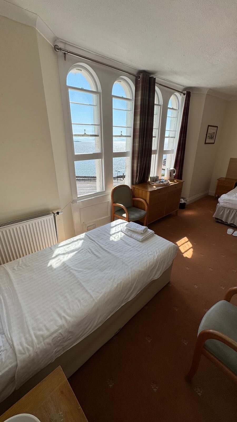 Habitación de hotel con una cama, una silla, un escritorio de madera y vista al mar a través de amplios ventanales.