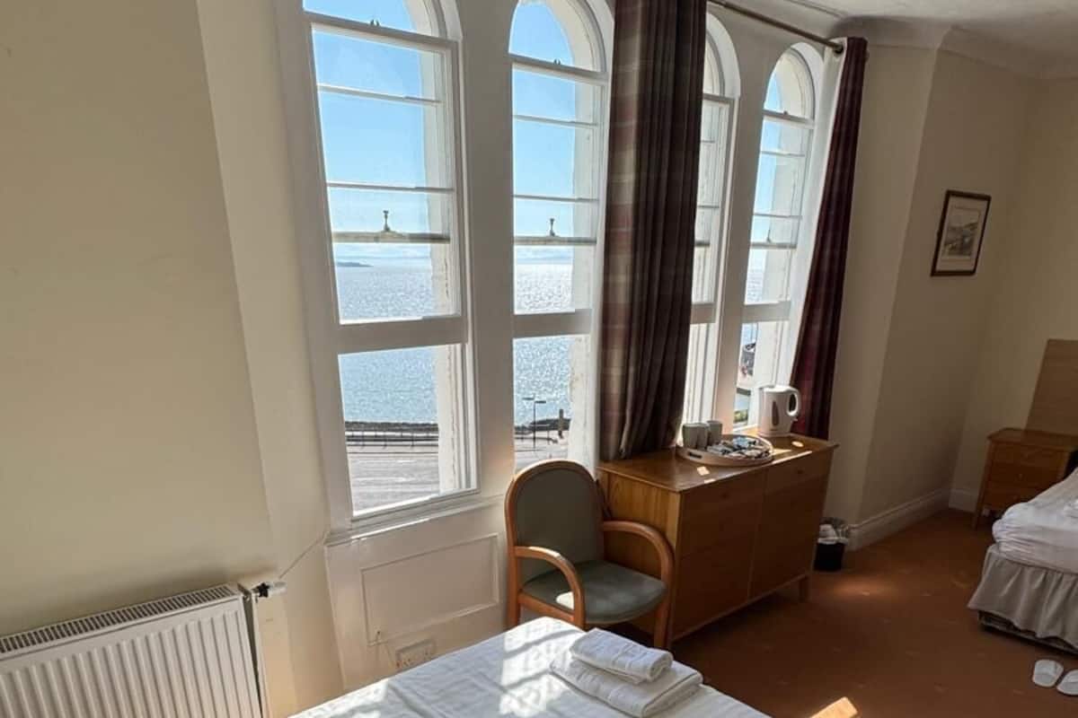 Une chambre d’hôtel avec un lit, une chaise, un bureau en bois et une vue sur la mer grâce à de grandes fenêtres.