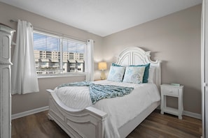 A bedroom with a large window, a white bed with a decorative headboard, a bedside table with a lamp, and a view of buildings outside.