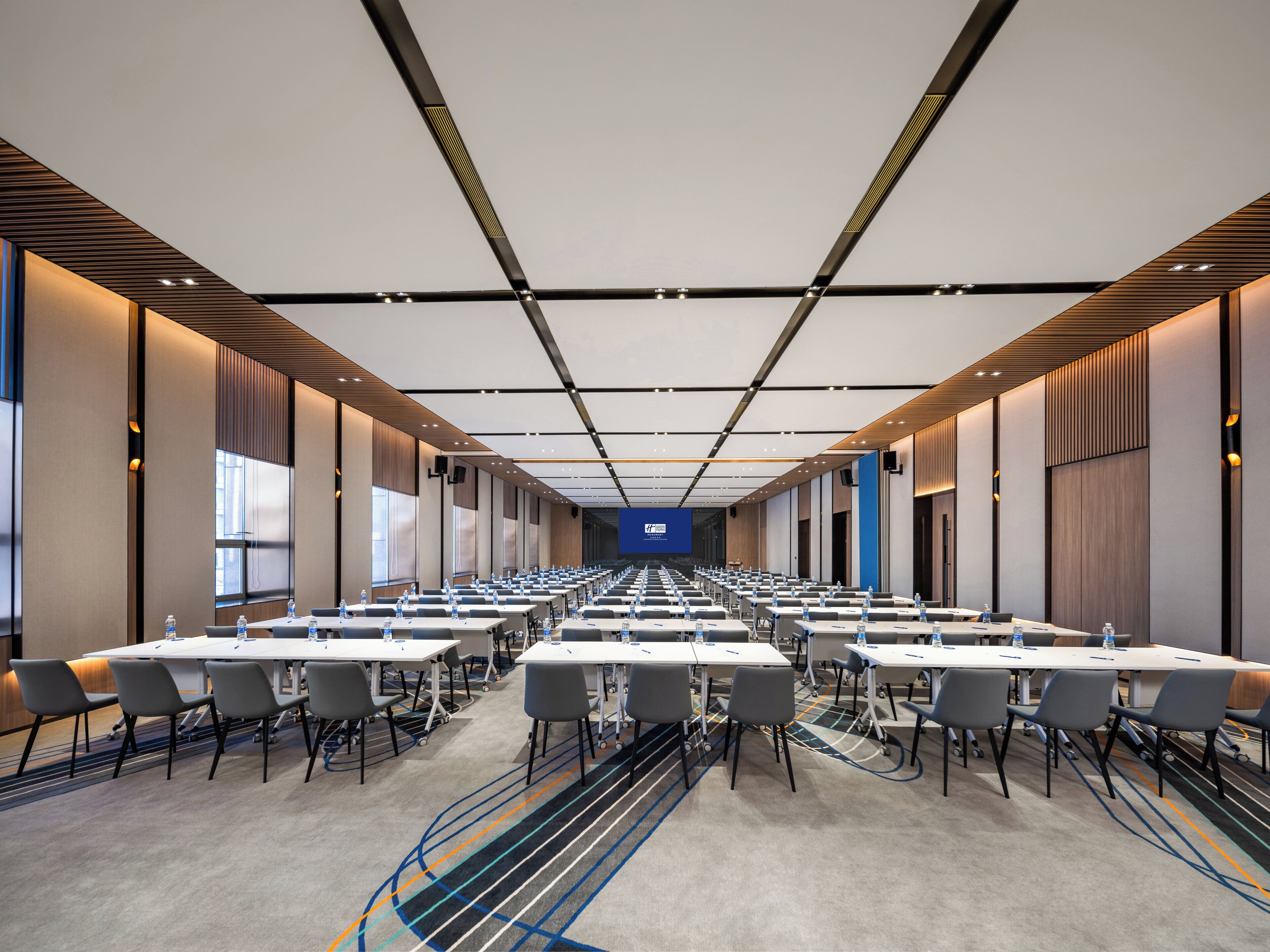 Una sala conferenze con file di tavoli e sedie, uno schermo grande nella parte anteriore e un moderno design del soffitto.