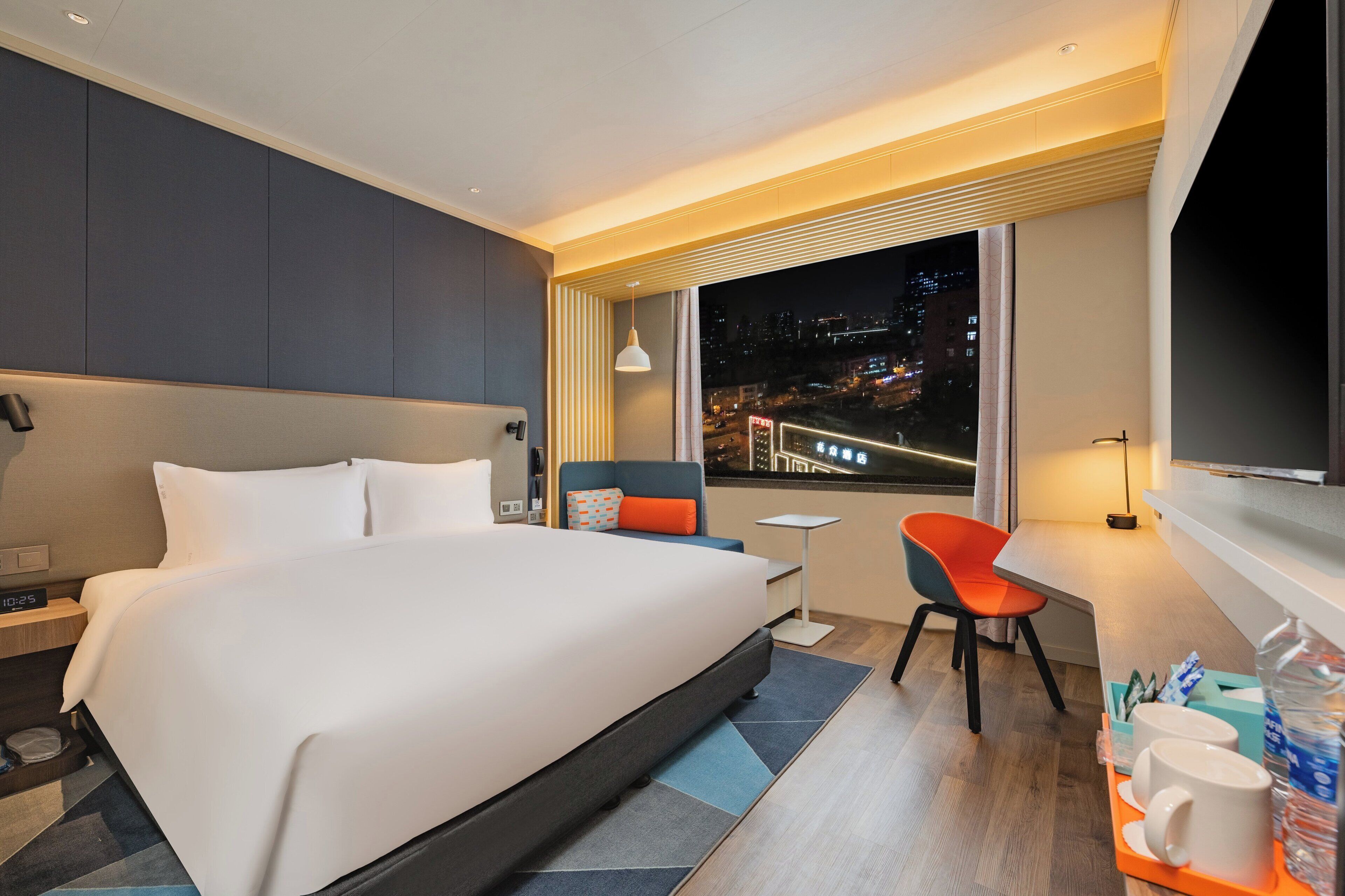 Habitación de hotel moderna con una cama grande, un escritorio, una silla y vistas a la ciudad de noche.
