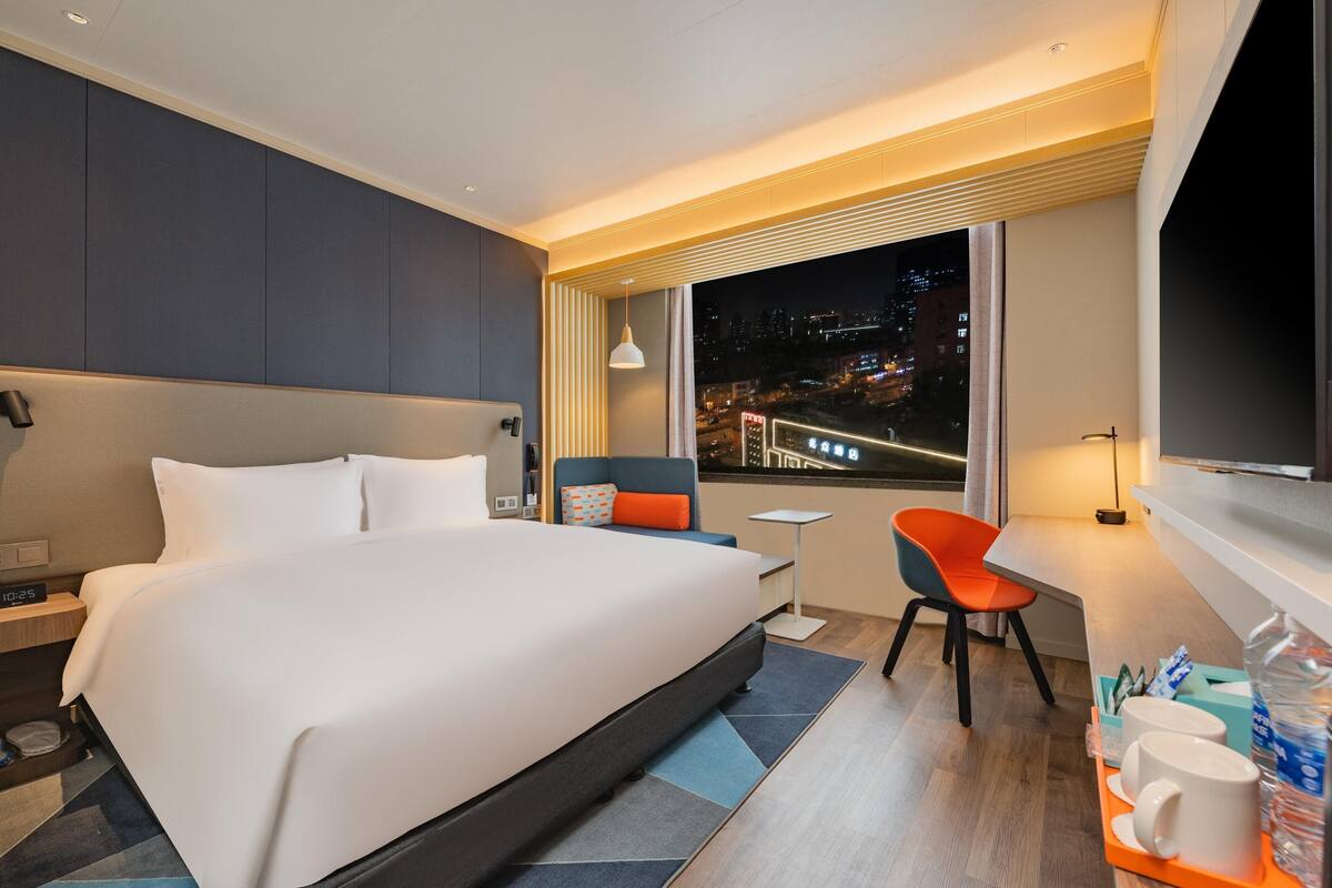 A modern hotel room with a large bed, a desk, a chair, and a view of the city at night.
