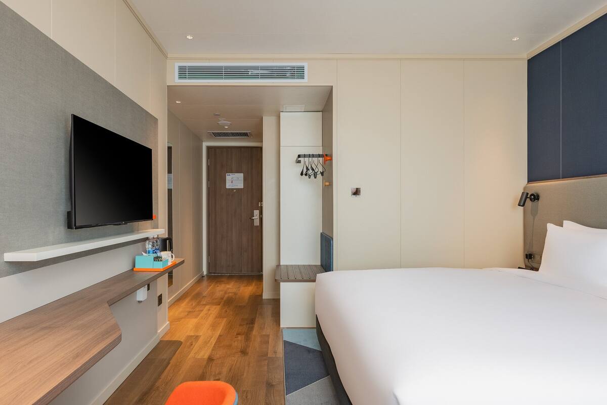 A hotel room with a bed, a TV mounted on the wall, a small table with a bottle, and a door leading to a bathroom.