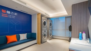 Ein moderner Waschraum mit blauer Wand, einer Sitzecke mit blauem Sofa und orangefarbenen Kissen sowie einer Reihe übereinander gestapelter Waschmaschinen.