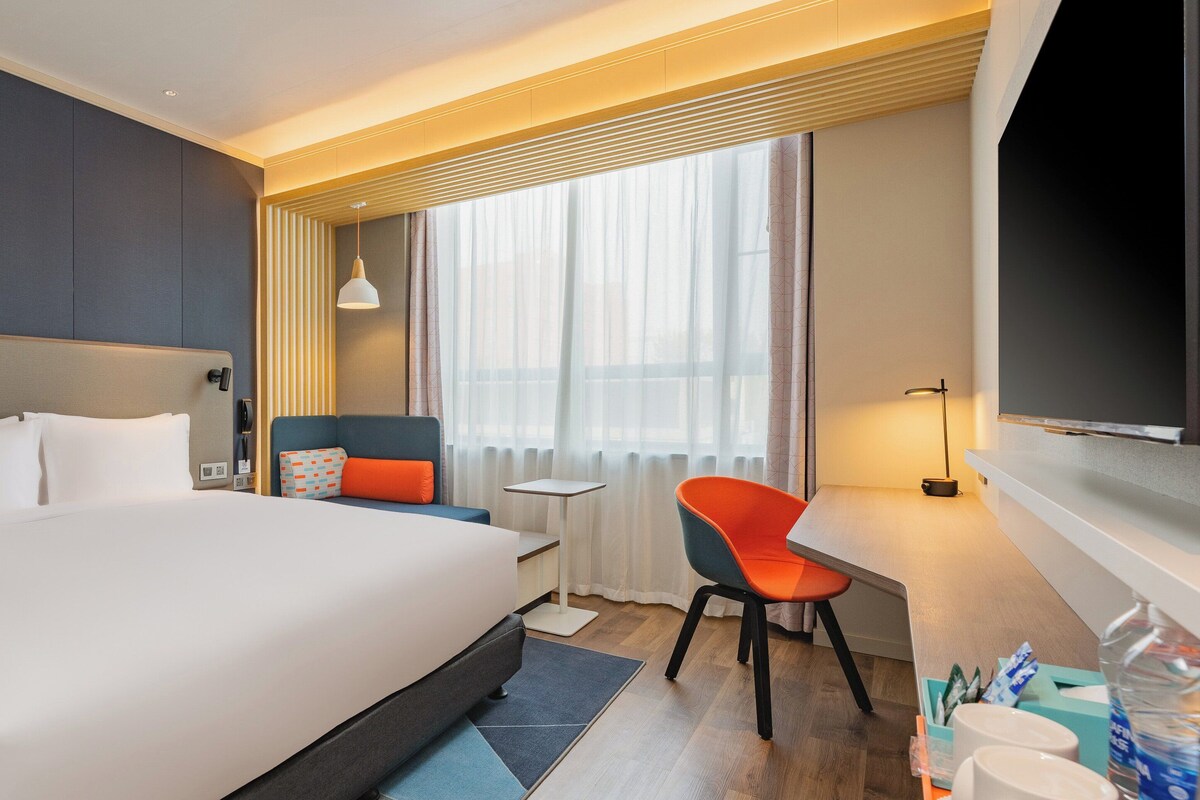 A modern hotel room with a large bed, a desk, a chair, and a TV.