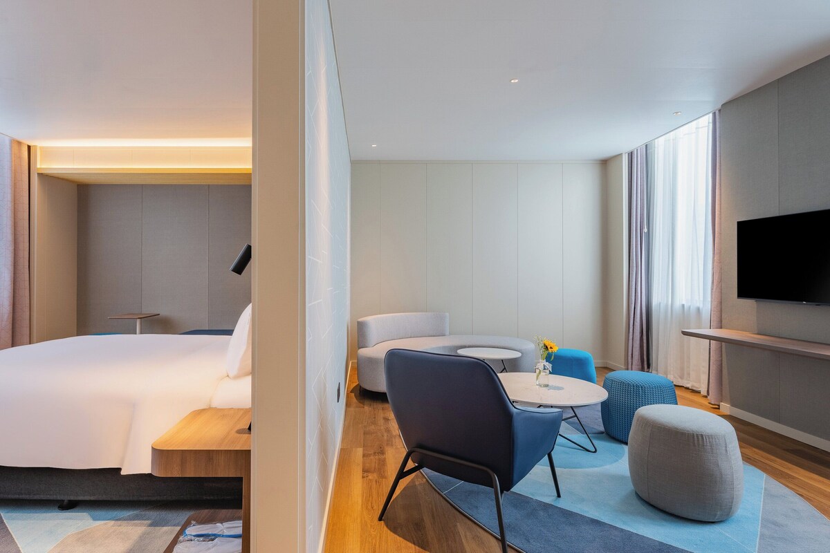 A modern hotel room with a large bed, a flat-screen TV, a sofa, a round coffee table, and ottomans.