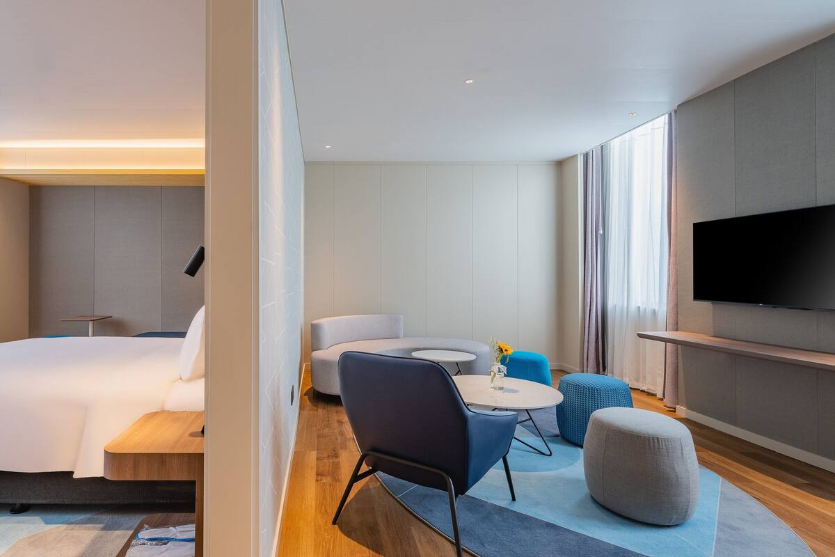 A modern hotel room with a large bed, a flat-screen TV, a sofa, a round coffee table, and ottomans.