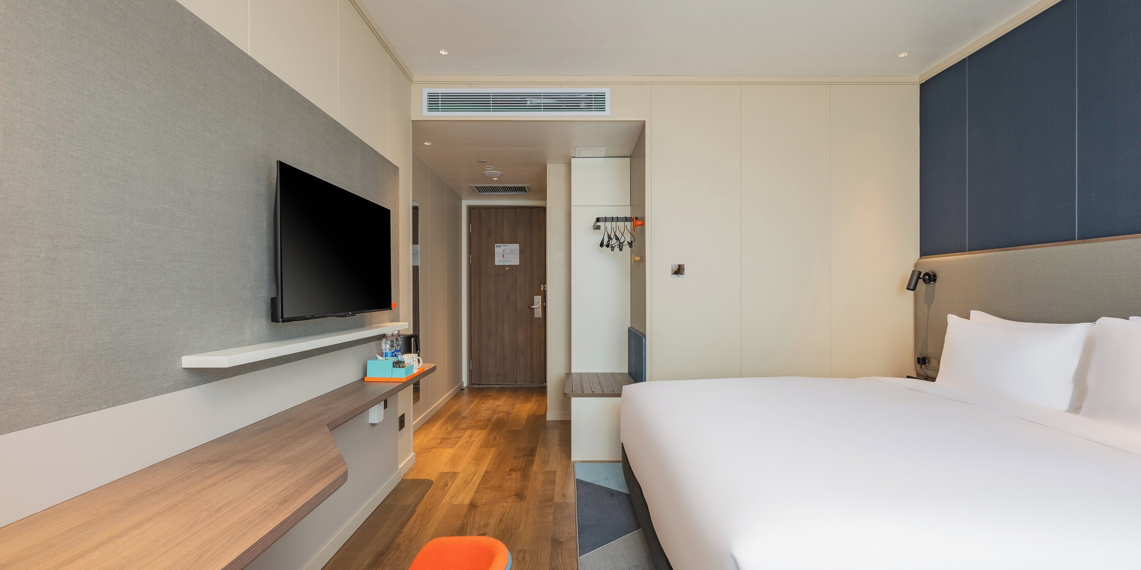 A hotel room with a bed, a TV mounted on the wall, a small table with a cup and a bottle, and a door leading to a bathroom.