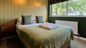 A bedroom with a patterned wallpaper, a bed with pillows, a nightstand, a window with a view of trees, and a radiator. - Pant Golau House (Llanbedr)