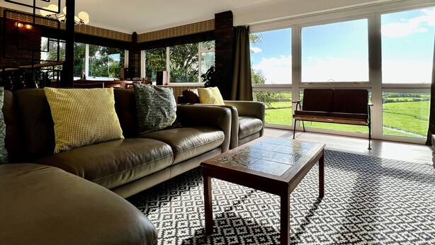 A spacious living room with a leather sofa, a patterned rug, and a view of the countryside through large windows. - Pant Golau House (Llanbedr)