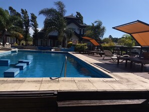 A swimming pool with steps leading into it, surrounded by lounge chairs and umbrellas, with a building in the background. - Complejo La Loma (Federacion)
