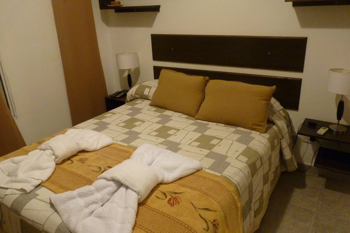 Una cama bien hecha con colcha a cuadros y dos almohadas, una mesita de noche con lámpara, y un estante en la pared sobre la cama.